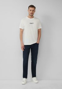 Man staat tegen een witte muur, gekleed in een witte T-shirt met logo, donkerblauwe jeans en witte sneakers, met een hand in de zak.