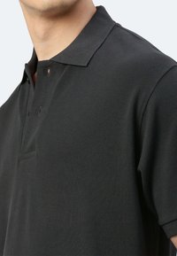 Schwarzes Poloshirt aus strukturiertem Stoff mit Kragen, zwei Knöpfen und kurzen Ärmeln. Deutliche Nähdetails betonen das Design.