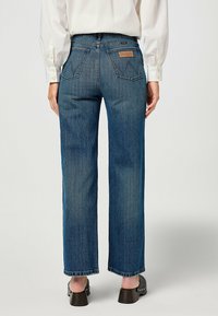 Jean denim bleu à jambes larges avec une taille mi-haute, présentant deux poches arrière ornées d'une couture en zigzag distinctive, ainsi qu'une étiquette de marque en cuir.