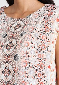 Haut à manches courtes imprimé floral avec un col rond. Principalement blanc avec des motifs orange, noir et gris. Tissu léger.