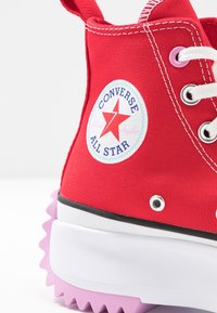 Baskets montantes en toile rouge avec lacets blancs, œillets violets et semelle violette en zigzag. Présente un logo circulaire Converse All Star.