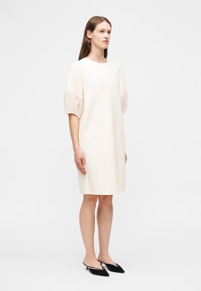 Γυναίκα που στέκεται φορώντας ένα άνετο, λευκό φόρεμα μέχρι το γόνατο με φουσκωτά κοντά μανίκια και μαύρα παπούτσια με μυτερή μύτη και χαμηλό τακούνι.