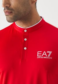 Polo rosso con tessuto testurizzato, colletto a tre bottoni e colletto bianco. Presenta il logo EA7 Emporio Armani sul lato sinistro del petto.