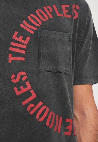 T-shirt cinza escuro feito de algodão com um design gráfico vermelho em texto redondo. Apresenta um bolso em patch no lado esquerdo do peito. Tecido macio e texturizado.