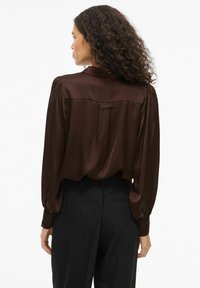 Blusa di satin marrone con maniche lunghe a sbuffo, dettaglio con bottoni ai polsini e cucitura sulla parte posteriore. Indossata con pantaloni neri su misura.