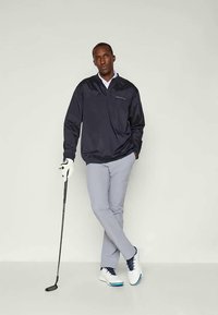 Tengerészeti pulóverdzseki, szürke nadrág és fehér golfcipő. A modell golfütőt tart, golfkesztyűt visel. Tiszta, minimális dizájn és modern fazon.