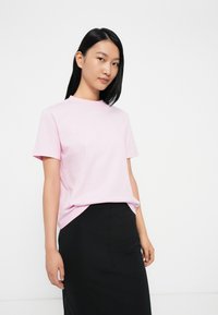 Ροζ βαμβακερό t-shirt με κοντά μανίκια και στρογγυλή λαιμόκοψη, φορεμένο πάνω από μια μαύρη φούστα. Απλές, καθαρές γραμμές και άνετη εφαρμογή.