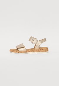 Gold metallic sandal with two wide straps, adjustable heel strap, and a circular decorative buckle. Cork-textured sole for added comfort.