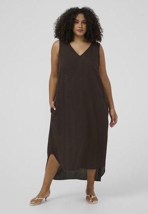 Göndör hajú nő sötétbarna, ujjatlan, V-nyakú midi ruhában és bézs színű, sarokkal ellátott szandálban, egyik kezét zsebre helyezve áll.