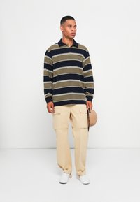 Gestreepte lange mouwen polo in olijf en marineblauw met witte strepen, beige cargobroek, witte sneakers, een beige pet in de hand. Minimalistisch ontwerp.