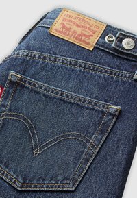 Les jeans en denim bleu de Levi's présentent une patch en cuir brun, des rivets en cuivre et un design de cœur cousu sur la poche arrière.