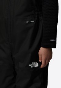Black waterproof jumpsuit featuring a zip front, adjustable waist, and breathable fabric. Includes the logos "DRYVENT" and "THE NORTH FACE."