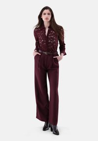 Burgunderfarbene Spitzenbluse mit Knopfleiste und Kragen, kombiniert mit hochtaillierten burgunderfarbenen weit geschnittenen Hosen. Schwarze Absatzstiefel runden den Look ab.