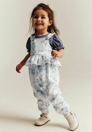 Lachende peuter met donker haar, gekleed in een blauw blousje met pofmouwtjes en witte bloemenprint tuinbroek, staand en lopend tegen een neutrale achtergrond.