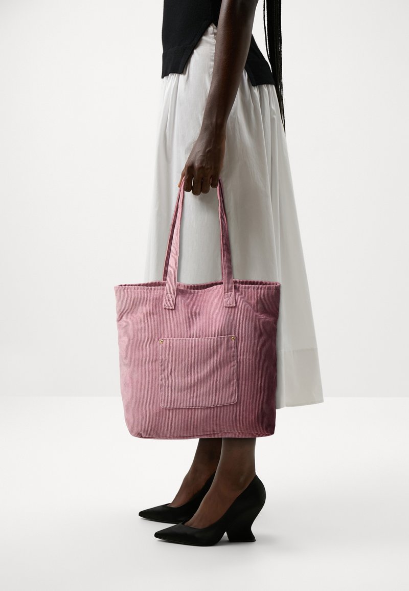 Personne portant une jupe blanche et un haut noir tenant un sac fourre-tout en velours côtelé rose, chaussée de talons noirs pointus sur fond blanc.