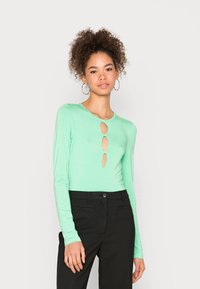 Blusa de manga longa verde menta com quatro recortes ovoides na frente, segurados por botões circulares, combinada com calças pretas ajustadas.
