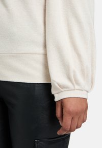 Cremefarbener, weich texturierter Pullover mit Puffärmeln, kombiniert mit schwarzen Lederhosen. Sichtbare Nahtdetails an der Taille und den Säumen.
