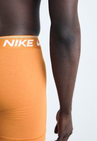 Nike oranžové boxerky s bielym logom a modrými detailmi. Hladká textúra s priliehavým strihom, zobrazujúca bočný pohľad na pás.