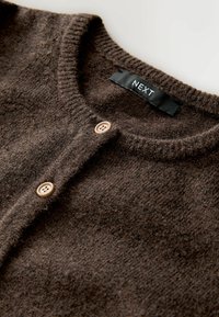 Brūns kardigāns, kas izgatavots no teksturēta auduma, ar apaļu kaklu, ribotām malām un divām gaiši brūnām pogām priekšpusē. Redzama zīmola etiķete.