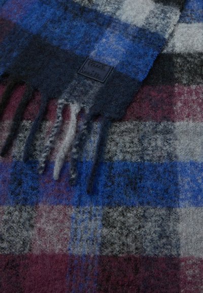 Vlněný šátek s kostkovaným vzorem v odstínech modré, šedé a vínové, s texturovaným povrchem a třásněmi na okrajích. Viditelný detail loga.