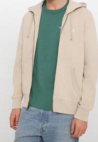 Sudadera beige con cremallera y capucha, con bolsillos frontales, superpuesta a una camiseta verde. Textura suave, corte casual, diseño minimalista, sin patrones visibles.