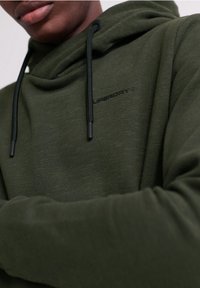 Felpa verde oliva realizzata in tessuto morbido, con cappuccio con cordino e un logo nero sul petto. Texture liscia e vestibilità casual.