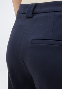 Παντελόνι σε ναυτικό μπλε με ραφή στην πλάτη και θηλιές για ζώνη, φορεμένο με μπεζ τοπ.