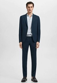 Completo blu navy con una vestibilità su misura, giacca strutturata e pantaloni slim. Abbinato a una polo azzurra e mocassini neri.