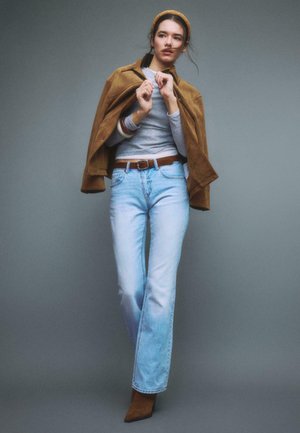 Junge Frau, die eine braune Jacke über die Schultern gelegt trägt, ein graues Langarmshirt, hellblaue Jeans und braune Stiefel, stehend vor einem grauen Hintergrund.