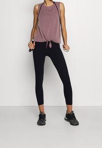 Femme en haut sans manches mauve et leggings noirs tenant une corde à sauter noire, debout sur un fond blanc uni.