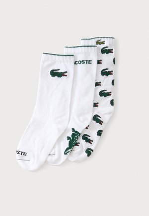 Tre hvide ankelsokker med grønne krokodillelogoer, en med gentaget logomønster og en med teksten "LACOSTE" på kanten.