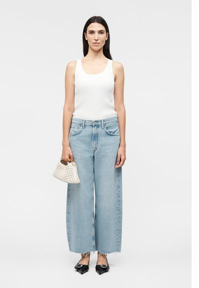 Γυναίκα στέκεται φορώντας λευκό τοπ, φαρδιά ανοιχτό μπλε τζιν, μαύρα μυτερά φλατ παπούτσια και κρατά μια μικρή λευκή πλεκτή τσάντα.