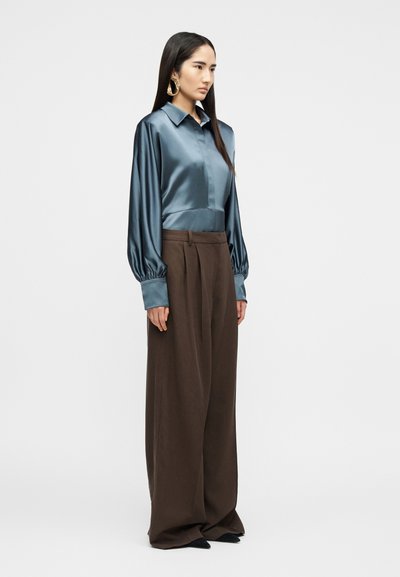 Femme portant un chemisier bleu brillant à manches longues et un pantalon large marron, debout de profil contre un fond clair uni.