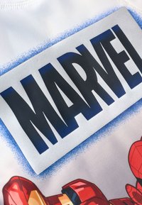 Tessuto bianco in poliestere con un grande e audace logo "MARVEL" nero su sfondo argento con accenti sfumati blu e illustrazioni di supereroi.