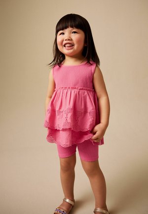 Young girl smiling, wearing a pink sleeveless dress with lace details and matching textured shorts, standing against a beige background.