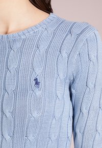 Vaaleansininen kabelikudottu neulepusero, jossa on ribbireuna ja pieni brodeerattu logo edessä. Rakennekuviointi, jossa on sileä viimeistely.