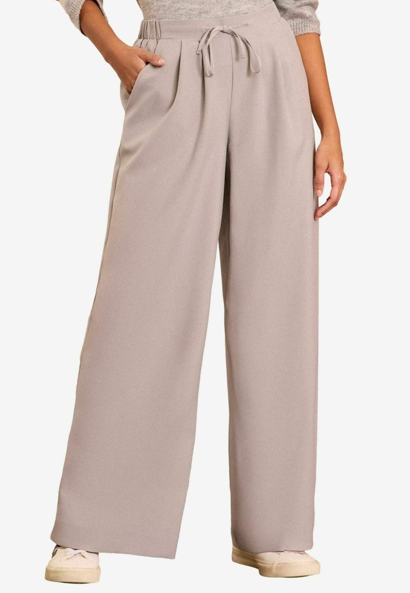 Široké nohavice z mäkkej béžovej tkaniny, s pásom na stiahnutie, záhybmi a bočnými vreckami, skombinované so svetlými teniskami.
