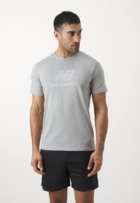 Svijetlosiva majica od mekog materijala s reljefnim logotipom New Balance u bijeloj boji. Uparen s crnim sportskim šortsevima.