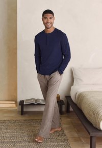 Navy long-sleeve henley shirt with three buttons, paired with plaid lounge pants in beige and brown tones; setting features a bed and rug.