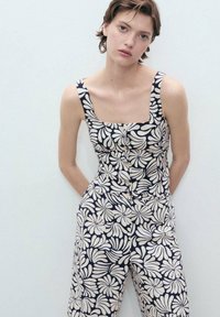 Navy floral jumpsuit featuring a square neckline, wide straps, and button front. The fabric has a textured finish with cream leaf patterns.