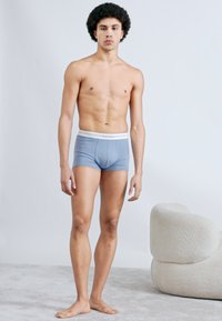 Boxers pour hommes bleus avec ceinture élastique blanche, tissu lisse, coupe ajustée. Modèle debout sur un sol gris clair.