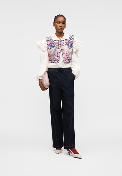 Femme portant un gilet brodé de fleurs sur une blouse blanche à volants, un pantalon noir à jambes larges, des talons blancs, et tenant une pochette rose.