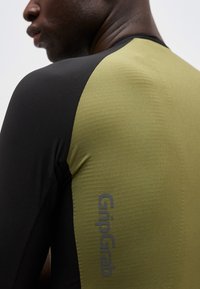 Κοντινό πλάνο ενός ατόμου που φοράει μακρυμάνικο μπλουζάκι σε μαύρο και ελιά πράσινο χρώμα με την επιγραφή "GripGrab" σε κάθετη διάταξη στην ελιά περιοχή.