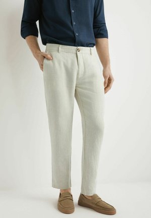 Mężczyzna ubrany w beżowe, dopasowane spodnie, granatową koszulę z długim rękawem i brązowe mokasyny, stojący z jedną ręką w kieszeni na białym tle.