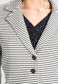 Bílý blazer s černými pruhy, se špičatým límcem a dvěma tmavými knoflíky. Nošený přes námořnické tričko s bílými puntíky.