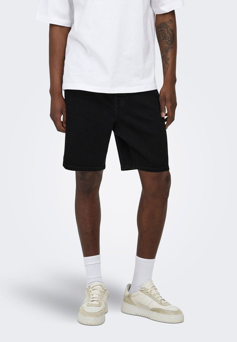 Personne portant un short noir, des baskets blanches avec des détails beige, des chaussettes blanches hautes et un t-shirt blanc à manches courtes avec un tatouage visible sur le bras.