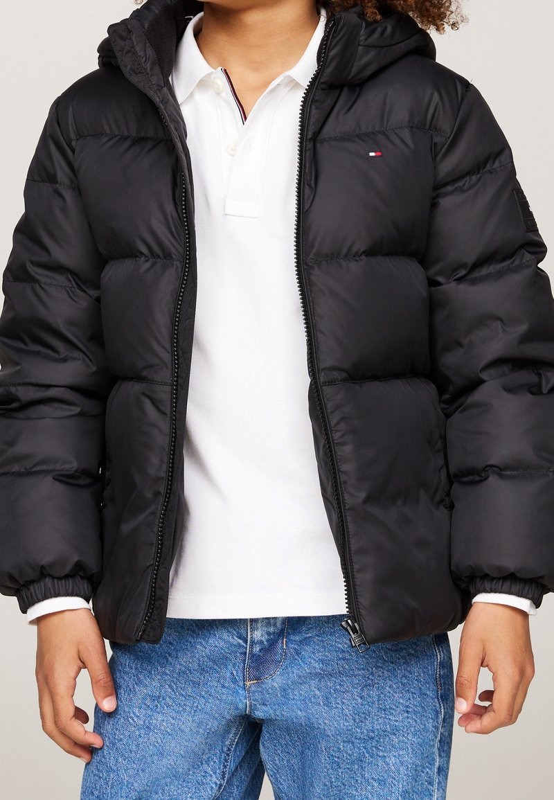 Schwarze Pufferjacke mit Kapuze, mit Reißverschluss vorne, gerippten Bündchen und Logodetail auf der Brust, getragen über einem weißen Poloshirt.