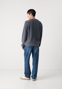 Szürke kötött pulóver bordázott részletekkel, kék farmernadrág felett viselve, fehér sneaker cipőkkel, hátulról nézve.