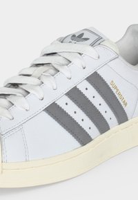 Λευκό αθλητικό παπούτσι Adidas Superstar με γκρι ρίγες, λευκά κορδόνια, χρυσή επιγραφή «SUPERSTAR» και εκρού σόλα σε ανοικτό φόντο.