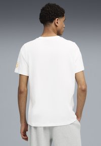 Camiseta blanca de manga corta con un cuello redondo y un logotipo amarillo en la manga izquierda. Material de algodón suave con un ajuste relajado.
